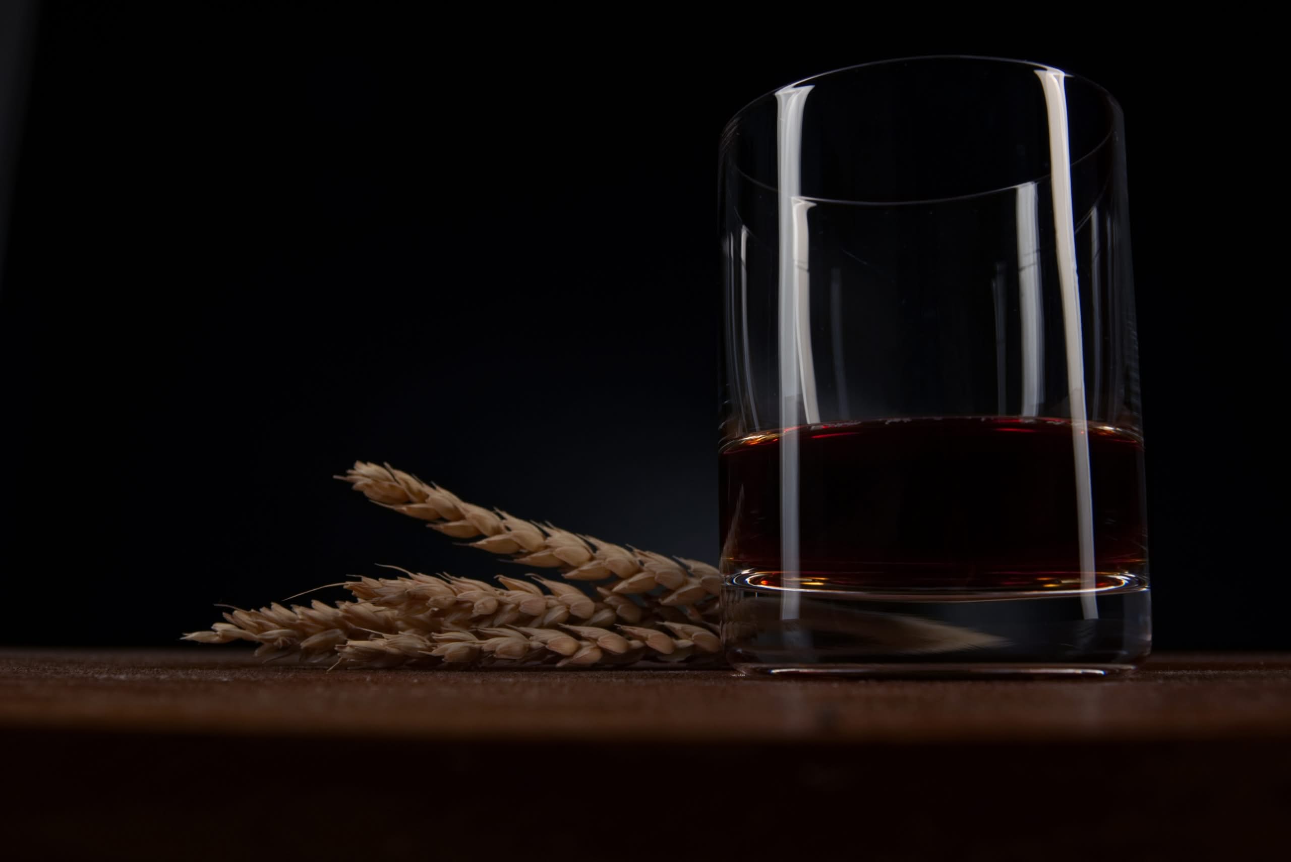 Bourbon whiskey in a rocks glass beside wheat stalks on a dark wooden surface