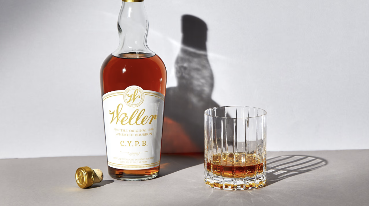 Weller C.Y.P.B. wheated bourbon bottle beside a glass of bourbon on a light grey surface